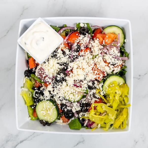 Overhead view of a vibrant Greek salad with feta, olives, cucumbers, tomatoes, and pepperoncini, with dressing.