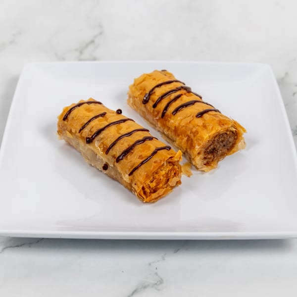 Two golden-brown baklava rolls drizzled with chocolate, on a white rectangular plate.
