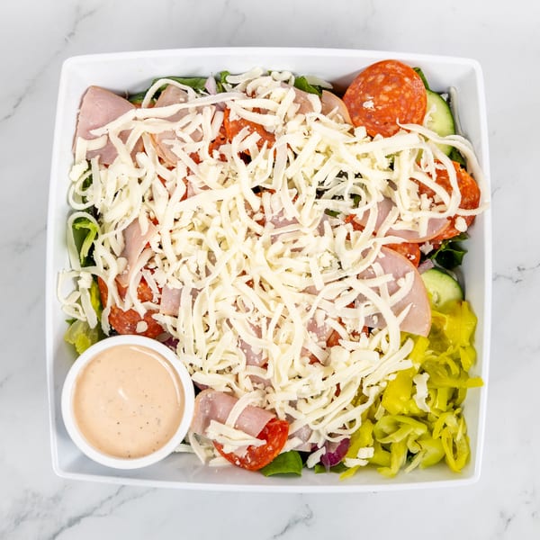 Overhead view of a large salad with ham, pepperoni, cheese, and peppers, with a side of dressing.