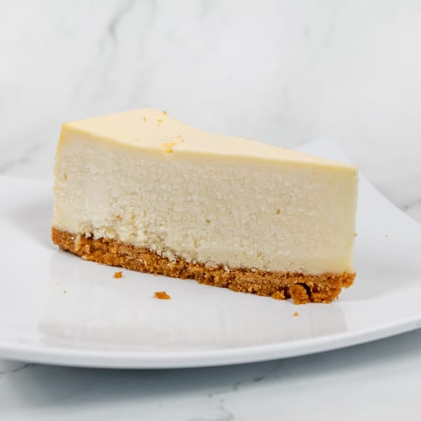 A slice of classic cheesecake with a graham cracker crust on a white plate.