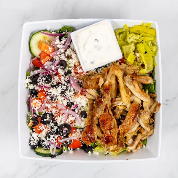 Overhead view of a white bowl filled with a Greek salad, chicken shawarma, and a side of tzatziki.