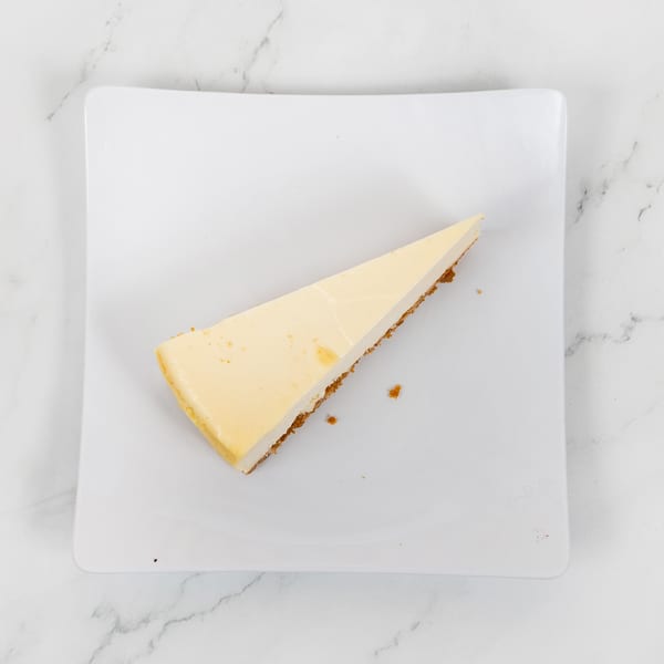 A slice of classic cheesecake with a graham cracker crust on a white square plate.