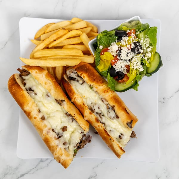 Two Philly cheesesteak sandwiches with fries and a side salad on a white plate.
